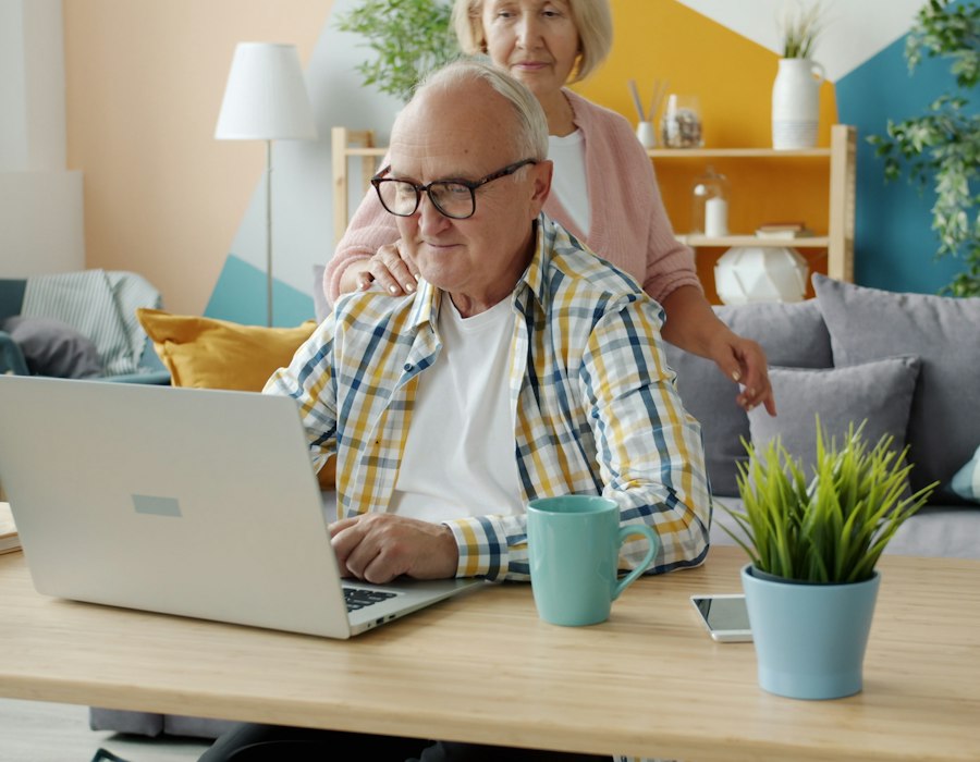 An older man at his laptop with his wife looking over his shoulder